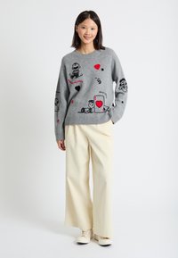 Grijze trui met cartoonprints in zwart en rood, met ribbelmanschetten en -zoom. Gecombineerd met ruime crèmekleurige broeken en beige schoenen.