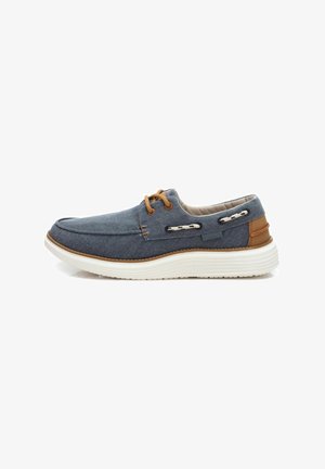 Zapato de lona azul marino con una suela blanca, acentos marrones y cordones decorativos; cuenta con una punta redondeada y un interior acolchado.