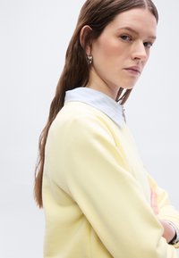 Νεαρή γυναίκα με μακριά καστανά μαλλιά, που φορά ασημένια κρίκους σκουλαρίκια, ανοιχτό μπλε πουκάμισο με γιακά κάτω από ένα ανοιχτό κίτρινο πουλόβερ, κοιτάζοντας πλάγια.