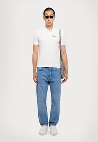 Hvit polo skjorte med logo, lyseblå denimjeans, grå crossover-veske og hvite joggesko. Modellen har på seg svarte solbriller mot en nøytral bakgrunn.