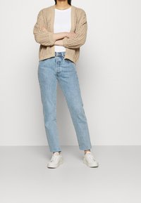 Cárdigan beige de punto con ajuste holgado, abertura frontal y patrón texturizado, combinado con vaqueros azul claro y zapatillas blancas.