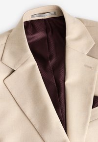 Blazer beige avec un col cranté et une doublure à motif bordeaux foncé, présenté à plat sur un fond blanc.
