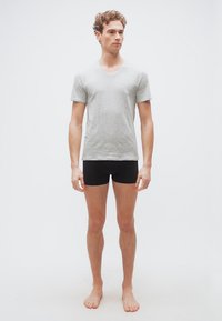 Jeune homme aux cheveux bouclés, pieds nus, portant un t-shirt gris clair à col en V et un boxer noir, se tenant devant un fond blanc uni.