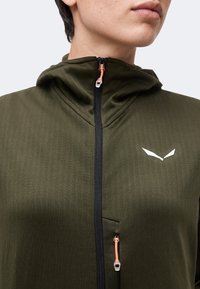 Κλείσιμο εικόνας ενός ατόμου που φοράει ένα ελιά-πράσινο μπουφάν με μαύρο φερμουάρ και πορτοκαλί λεπτομέρειες φερμουάρ, δείχνοντας ένα λευκό λογότυπο σε σχήμα πουλιού στο στήθος.