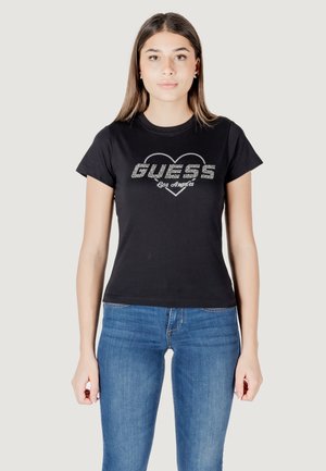 T-shirt nera in cotone a maniche corte, con un grafico a forma di cuore e il testo glitterato "GUESS Los Angeles" sul davanti. Indossata con jeans blu.