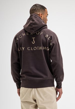 Man draagt een donkerbruine hoodie met gedeeltelijk verborgen beige tekst op de rug en lichtbeige broek, staand en met de rug naar voren.