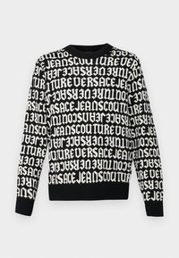 Maglione nero lavorato a maglia con testo bianco che ripete "VERSACE JEANS COUTURE" in varie orientazioni. Collo e polsini a costine.