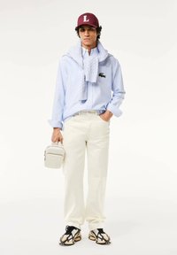 Camisa de botones a rayas azul claro, suéter de punto blanco colgado, pantalones crema, zapatillas de colores, gorra burdeos con "L", llevando un pequeño bolso crema.