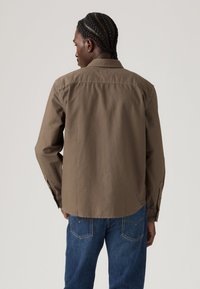 Persoon met gevlochten haar draagt een bruine lange mouwen shirt en blauwe jeans, met de rug naar de camera tegen een effen lichte achtergrond.