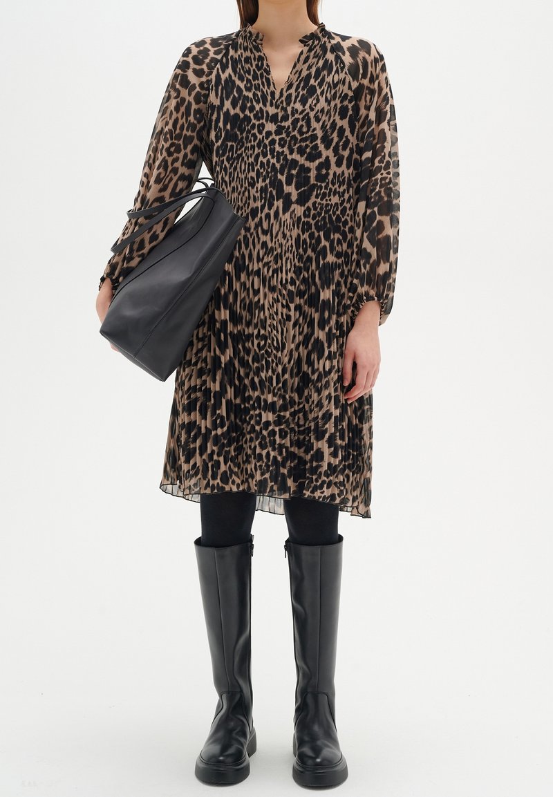 Femme portant une robe plissée à imprimé léopard, des collants noirs, des bottes noires montantes jusqu'au genou, tenant un grand sac à main en cuir noir.