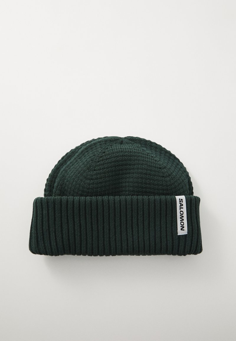 Dark green knitted beanie with ribbed texture, featuring a folded cuff and a white logo tag sewn on the side.