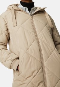 Chaqueta acolchada beige con capucha, que presenta costuras diagonales, cierre de cremallera y puños acanalados. Los bolsillos laterales están integrados en el diseño.