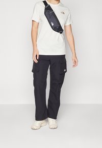 Mustat cargo-housut, joissa on useita taskuja, vaaleanbeige t-paita ja pieni musta olkalaukku. Jalkineet ovat kermanväriset tennarit.