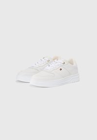 Une paire de baskets basses blanches avec des détails perforés, des lacets blancs et un petit logo rectangulaire sur le côté, présentées sur un fond uni.
