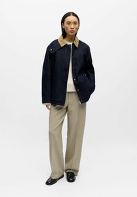 Giacca di jeans blu scuro con colletto color cuoio, tasche frontali e chiusure a scatto. Abbinata a pantaloni beige e scarpe nere. Design minimalista.