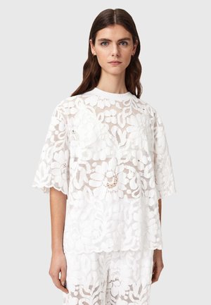 Vrouw draagt een witte top met korte mouwen van bloemenkant en bijpassende broek tegen een effen lichtgrijze achtergrond.