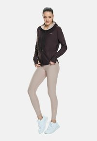 Žena stoji, odjevena u tamni hoodie sa zatvaračem i rupama za palčeve, bež tajice i bijele tenisice, nasuprot jednostavnoj bijeloj pozadini.