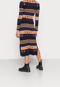 Robe en maille avec des rayures navy, noir et beige et un motif de texte "LOVE". Manches longues, fentes latérales, portée avec des bottines noires.