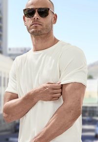 Bílý tričko s krátkým rukávem, které nosí osoba s krátkým účesem, doplněné brýlemi ve stylu želvoviny, ukazující hladkou strukturu látky.