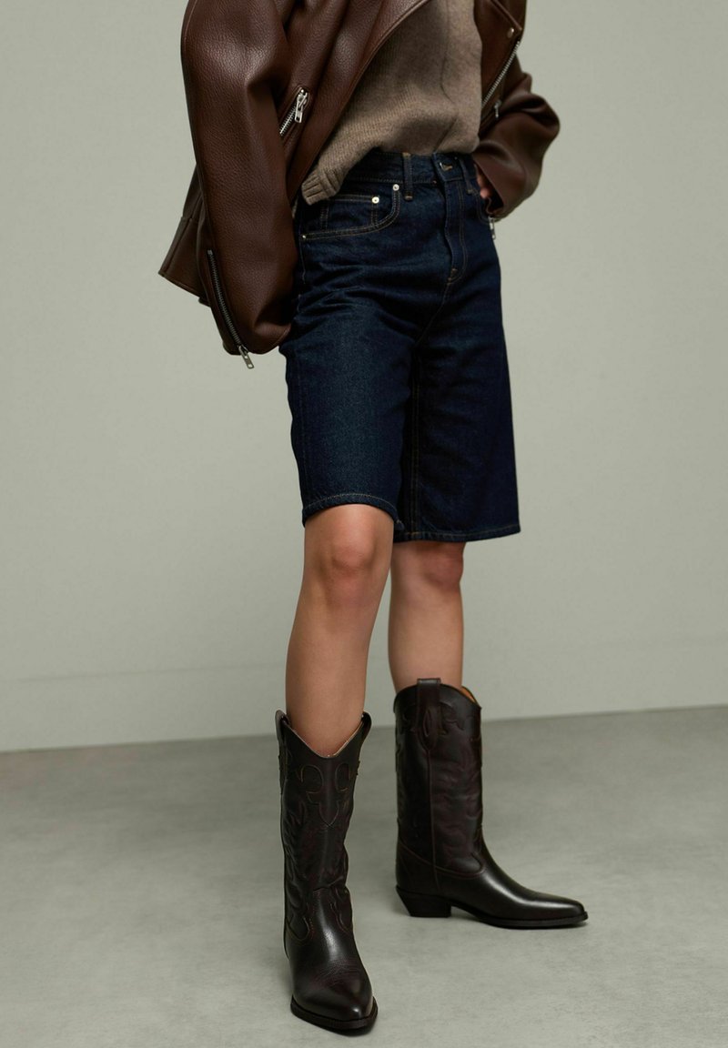 Bermuda en denim bleu foncé avec une coupe droite, associée à des bottes de cowboy en cuir marron ornées de coutures décoratives et à bout pointu.