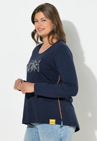 Camicia a maniche lunghe blu navy con grafica bianca, presenta un accento arancione sulla cucitura laterale e un'etichetta logo gialla. Tessuto morbido e strutturato.