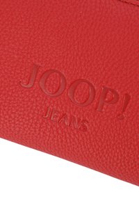 Rdeča usnjena denarnica z teksturo, ki prikazuje vtisnjen logotip "JOOP! JEANS". Površina ima grobo teksturo, kar dodaja globino oblikovanju.