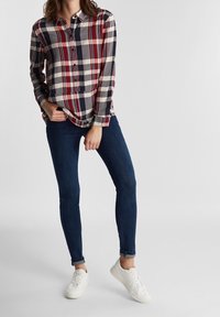 Donna che indossa una camicia a quadri rossa, blu navy e bianca, jeans skinny blu scuro e sneakers bianche, in piedi contro uno sfondo neutro.