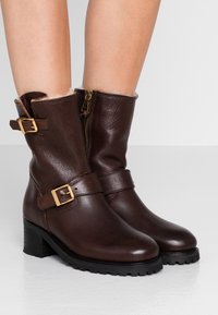Hnědé kožené kotníkové boty s texturovaným povrchem, s jazýčky zlaté barvy a bočním zipem. Patkový podpatek a polstrovaný límec pro pohodlí.