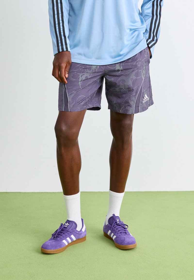 Henkilö, joka pitää yllä purppuroita Adidas-shortsseja, vaaleansinistä pitkähihasta toppia, valkoisia sukkia ja purppuroita Adidas-tennareita, seisoo vihreällä lattialla.