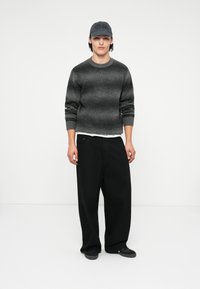 Γκρι ριγέ πουλόβερ με ribbed μανσέτες, φορεμένο πάνω από λευκό πουκάμισο και συνδυασμένο με χαλαρές μαύρες παντελόνες και σκούρα αθλητικά παπούτσια, φέροντας ένα γκρι καπέλο.