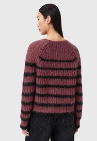 Femme aux longs cheveux foncés portant un pull violet côtelé avec des rayures horizontales noires et un pantalon noir, vue de dos.