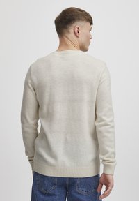 Suéter de punto beige claro con cuello redondo, puños y dobladillo acanalados, con un acabado texturizado, usado sobre jeans de denim azul.
