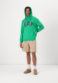 Felpa verde con logo "GAP" in blu navy, tasca a marsupio e cappuccio con cordino. Abbinata a pantaloncini beige e sneakers color sabbia. Calzini bianchi visibili.