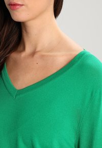 Pull vert à col en V avec une texture douce, présentant une coupe décontractée et des épaules légèrement tombantes. Tissu lisse sans motifs distinctifs.