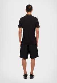 Čierna polo košeľa s vzorovaným golierom a manžetami, v kombinácii s čiernymi šortkami a čiernymi topánkami, z pohľadu zozadu.