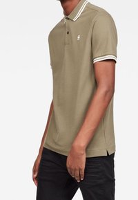 Polo shirt vert olive avec un col, manches courtes avec des rayures blanches, et un petit logo sur la poitrine. Texture en coton lisse.