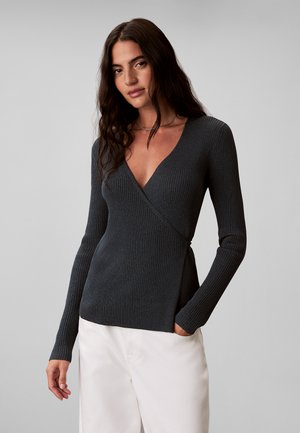 Vrouw met lang donker haar die een donkergrijze geribde omslagtrui en witte broek draagt, staand met één hand in de zak.