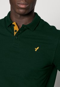 Dunkelgrünes Polohemd mit kurzen Ärmeln, das einen gelben Kragen und ein kleines gelbes gesticktes Vogel-Logo auf der Brust hat.