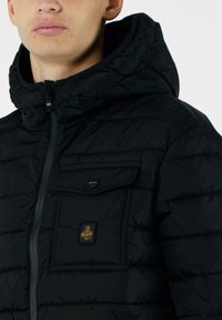 Giacca a vento nera con cappuccio, con zip frontale, texture trapuntata e tasca sul petto. Include una patch con il logo sulla tasca.