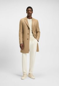 Cappotto lungo beige in lana con rever a scollo, chiusura con bottoni, abbinato a un maglione crema strutturato e pantaloni bianchi su misura, insieme a sneaker chiare.