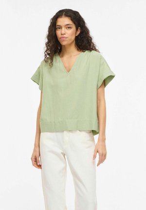 Kobieta z kręconymi włosami, ubrana w luźny, jasnooliwkowy top z krótkim rękawem i dekoltem w serek oraz białe spodnie, stojąca na tle jednolitego tła.