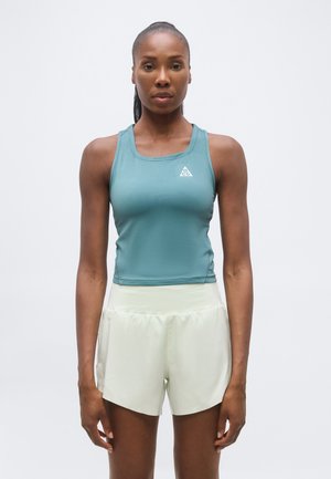 Γυναίκα που στέκεται με το σώμα στραμμένο προς τα εμπρός, φοράει ένα αμάνικο αθλητικό τοπ σε τιρκουάζ χρώμα και ανοιχτό μπεζ σορτς, με φόντο μια απλή γκρι επιφάνεια.