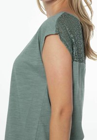Teal kortærmet top med tekstureret blonderdetail på skuldrene. Stoffet virker blødt med en afslappet pasform og enkelt design.