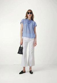 Nainen, jolla on mustat aurinkolasit, vaaleansininen lyhythihainen paita, valkoiset capri-housut ja mustat liukuva kengät, pitää kädessään pientä mustaa käsilaukkua.