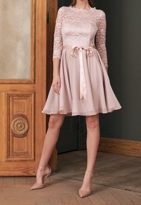 Woman wearing a pale pink lace top dress with three-quarter sleeves, ribbon belt, and flared skirt, paired with nude high-heeled shoes.