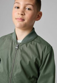 Roheline pommitaja jakk, millel on lukuga eesmine osa, soonikkoes krae ja diskreetne logo tikand rinnal. Sile, kergesti kantav kangas.