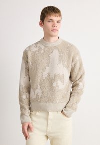 Young man with short hair wearing beige and cream textured sweater and light cream pants, standing against a plain white background.