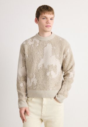 Young man with short hair wearing beige and cream textured sweater and light cream pants, standing against a plain white background.