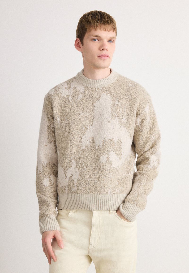 Young man with short hair wearing beige and cream textured sweater and light cream pants, standing against a plain white background.