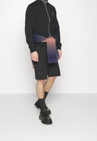 Personne portant une veste zippée noire, un short noir, des bottes noires épaisses et une écharpe dégradée violet-rose nouée autour de la taille.